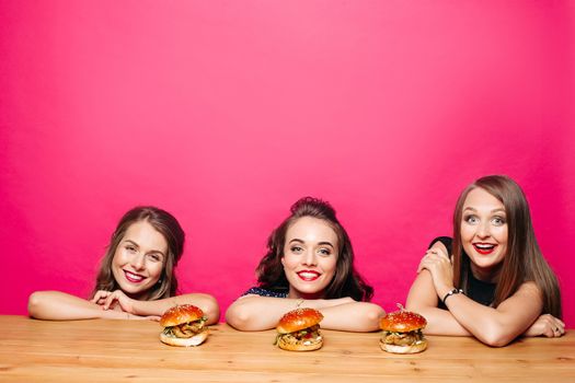 Studio portrait of beautiful ladies smiling happily at camera sitting at burgers on wooden table before eating them. Isolate on pink background. Copyspace. Advertising.