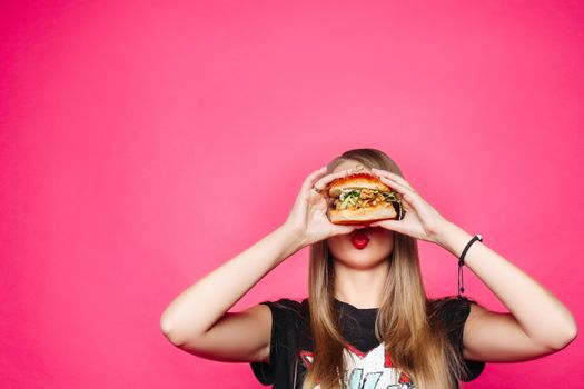 Close-up of pretty long-haired girl biting delicious burger with chicken and salad, looking at camera against pink background. American fast food concept.