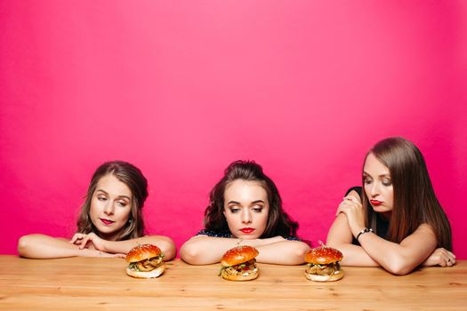 Portrait of three girlfriends with make up looking sadly and pensively on three delicious homemade burgers on wooden table. Isolate on pink background.
