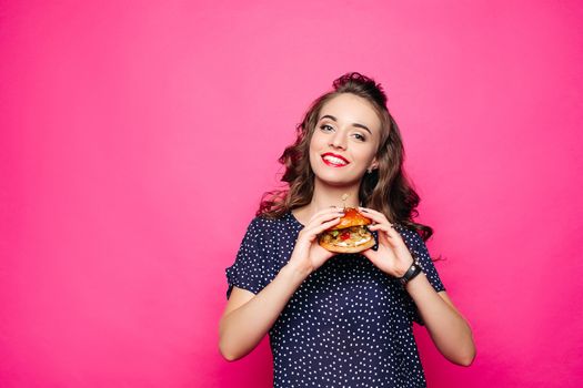 Happy girl holding a hamburger. Happy offers to try a sandwich, looking apetitnym with a look at you. The student is eating a hamburger. Isolated on a pink background
