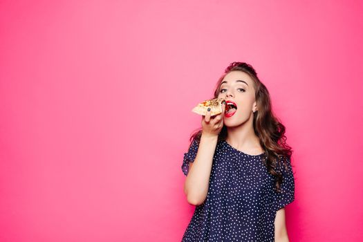 Happy girl eating a pizza. She opened her mouth, holding a pizza in her right hand and staring at you with satisfaction. Isolated on pink background