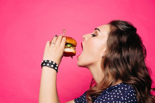 Close up of hungry girl with opened mouth, holding and eating big hamburger. Pretty woman with curly hairstyle and red lips eating tasty cheeseburger with tomato and meal. Concept of fast food.