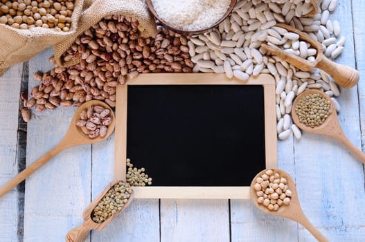 Different types of beans around black framed square on blue wooden table. From above.