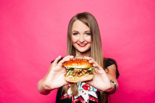 Studio portrait of beautiful happy lady with long fair hair holding cheeseburger with chicken and salad in hands and smiling at camera over pink background.