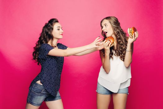 Two pretty and young hungry hipster girls with opened mouth, giving, holding and wanting two big hamburger. Best girlfriend with long hair, make up hiding tasty cheeseburger with tomato and meal.