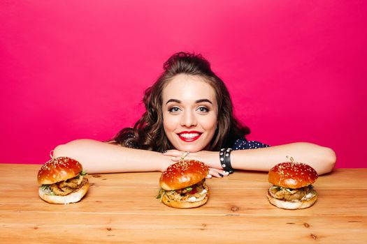 Portrait of excited woman with brunette hair smiling at camera leaning on wooden table with three delicious burgers with chicken.