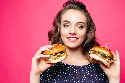 Beautiful positivity girl smilingat camera and posing with two tasty humburgers. Young pretty woman wearing in black holding big cheeseburgers. Pink studio background. Concept of fast food and diet.