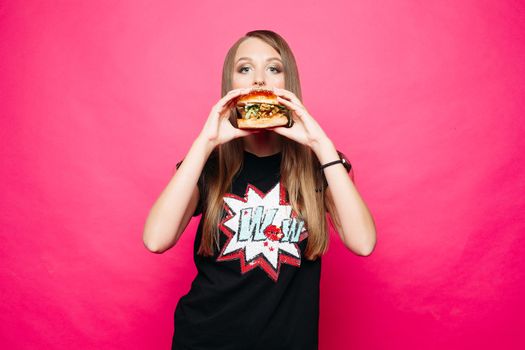 Studio portrait of fair haired girl in black t-shirt with print eating hamburger and looking at camera against vivid magenta background.