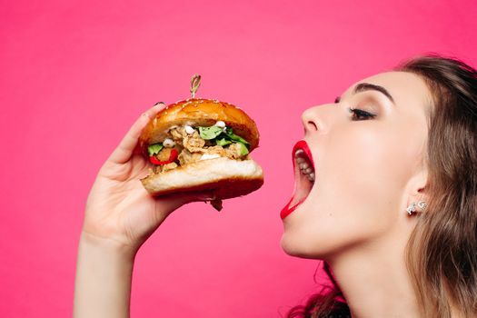 Close up of hungry girl with opened mouth, holding and eating big hamburger. Pretty woman with curly hairstyle and red lips eating tasty cheeseburger with tomato and meal. Concept of fast food.