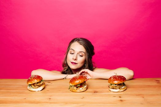 Portrait of unhappy pretty girl with hairstyle and make up looking at delicious freshly cooked hamburgers on wooden table with her chin on hands over bright magenta background.