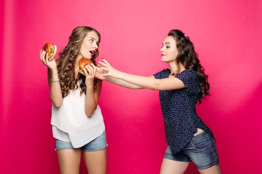 Two pretty and young hungry hipster girls with opened mouth, giving, holding and wanting two big hamburger. Best girlfriend with long hair, make up hiding tasty cheeseburger with tomato and meal.