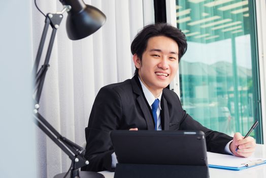 Work from home, Asian young businessman video conference call or facetime he smiling looking to camera sitting on desk using smart digital tablet computer and write notebook information at home office