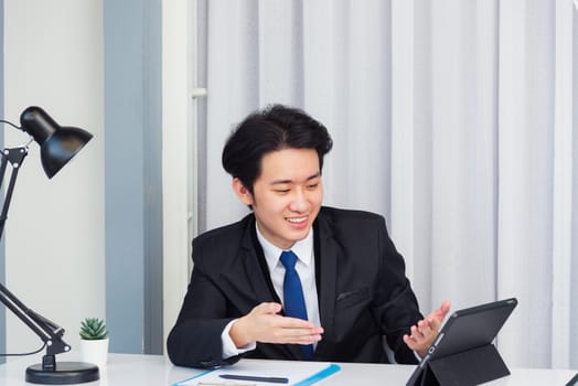 Work from home, Asian young businessman video conference call or facetime he smiling looking to tablet sitting on desk using smart tablet computer raise hand and explain to colleagues at home office