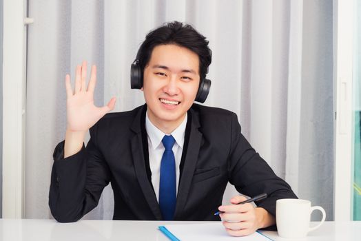Work from home, Asian young businessman wear suit video conference call or facetime he smiling sitting on desk wearing headphones and raise your hand to say hello greet colleague at home office