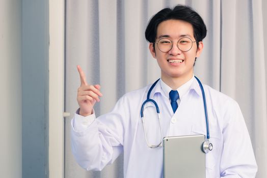 Portrait closeup of Happy Asian young doctor handsome man smiling in uniform and stethoscope neck strap holding smart digital tablet on hand and point finger to side away, healthcare medicine concept