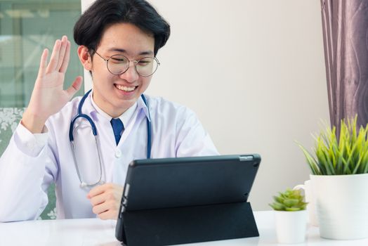 Happy Asian young doctor handsome man smile in uniform wear glasses raise hands to greet patients modern smart digital tablet computer on desk at hospital office, Technology Healthcare And Medicine