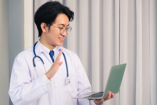 Asian young handsome doctor man wearing a doctor's dress and stethoscope video conference call or facetime raise hand to say hello he smiling at hospital office, Health medical care concept