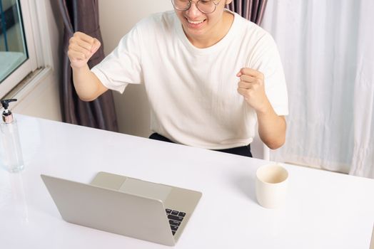 Happy Asian young business handsome man smile work from home office wear glasses, t-shirt comfortable exciting good news successful winner raises hand yes gesture with laptop computer on desk at home