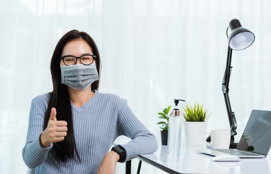 Asian young business beautiful woman wearing face mask protective working from home office with laptop computer desk he quarantines disease coronavirus her show finger thumber for good job sign