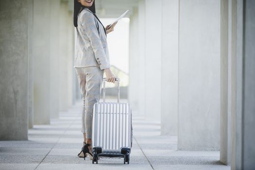 Portrait of business woman looking digital tablet with white travel bag on walkway while waiting to travel to the destination 