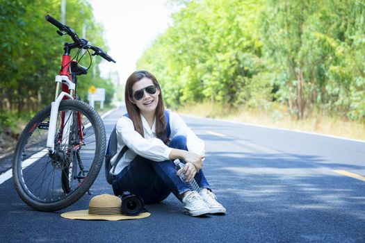 Beautiful woman sat on the side of the bicycle after biking to travel along the paved road on both sides of the road, full of beautiful green trees. The concept of travel and freedom.