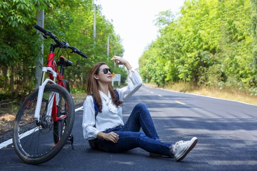 Beautiful woman sat on the side of the bicycle after biking to travel along the paved road on both sides of the road, full of beautiful green trees. The concept of travel and freedom.