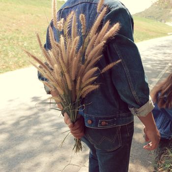 The man hides the grass flowers behind his back to surprise his girlfriend.