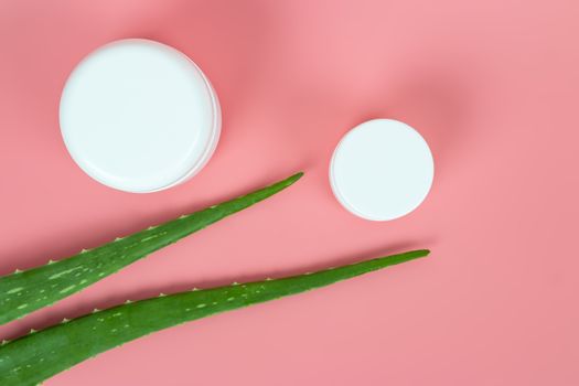 Fresh aloe vera leaves and white plastic cosmetic jar on pastel pink background for Health and beauty products.