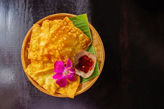 Yellow fried dumplings and a cup of sauce on a bamboo dish at decorated with orchid placed on the table dark wood. Select focus.