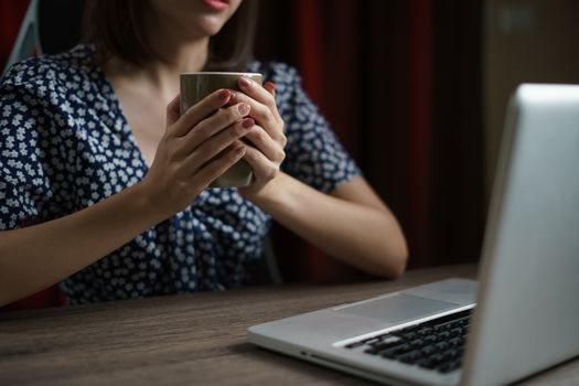 Woman working from home using computer and drinking cup of tea, closeup portrait indoor.