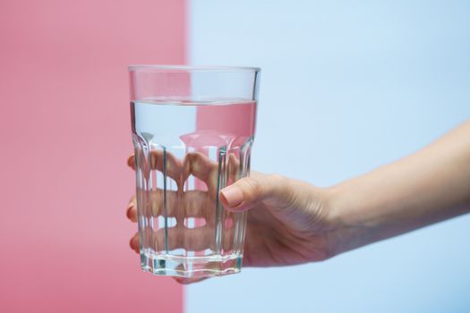 Hand holding transparent glass of water, clean drinking water in a clear glass on two tone (pink and blue) background