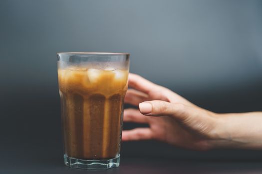 Hand with ice coffee in a tall glass on dark background.