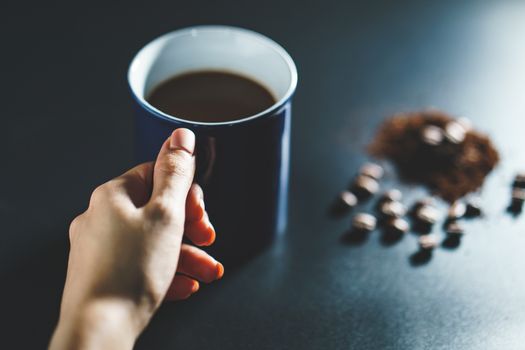 Hand holding a cup of coffee with coffee beans and powder on black background.