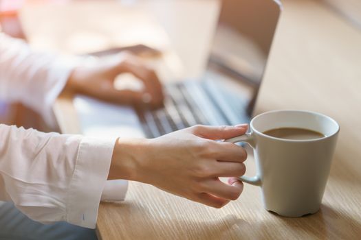 Woman hand on work desk with a laptop computer, a cup of coffee, with morning sunlight.