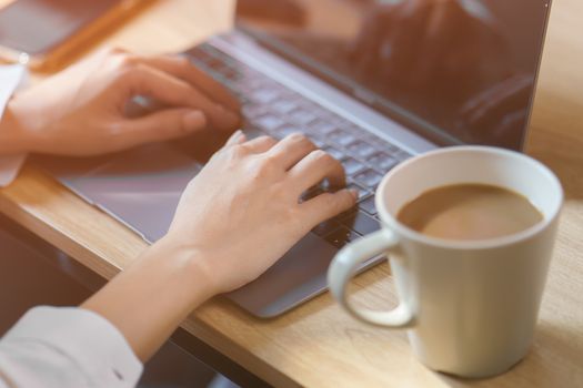 Woman hand on work desk with a laptop computer, a cup of coffee, with morning sunlight.