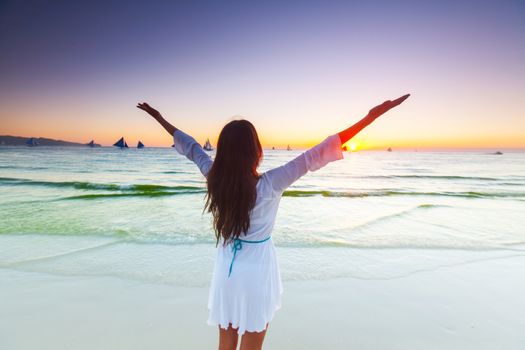 Woman standing on tropical sea beach with raised arms up and looking into a wide sea view with sail boats