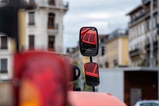 Close-up of tractor side mirror on defocused city background. The mirror shows the red hood of the tractor - image