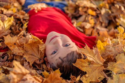 little boy in red looks at the camera and smiles while lying in the fallen yellow leaves in the autumn park