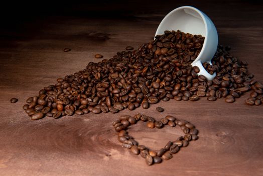 Coffee bean pouring out of a white mug on wooden table, Heart shape from coffee beans in the foreground - image