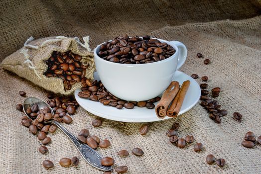 White coffee mug full of organic coffee beans and  cinnamon sticks on linen cloth - image