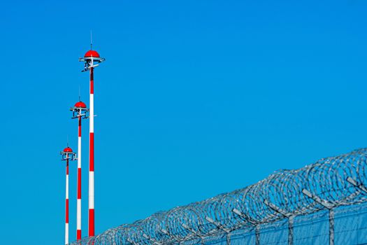 fence with barbed wire and row of the airport lamp posts with alternate red and white painting against blue sky, security concept