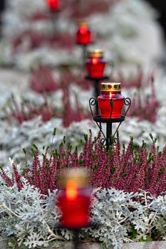 row of burning candles in red glass holders on a defocused floral background at night, remembrance event