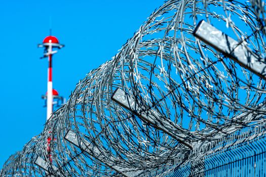 fence with barbed wire and row of the airport lamp posts with alternate red and white painting against blue sky, security concept