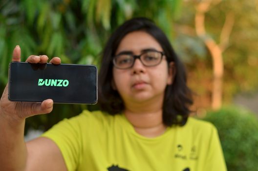 Mumbai, India, 2020. Young Girl with Dunzo Delivery app logo glowing on the mobile phone screen in her hands. Selective focus and shallow depth of field. The main focus is on the logo.