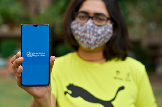 Geneva, Switzerland,2020. Girl wearing mask showing on her mobile phone screen, World health organization (WHO) landing page & emblem.Specialized agency of United Nations responsible for public health