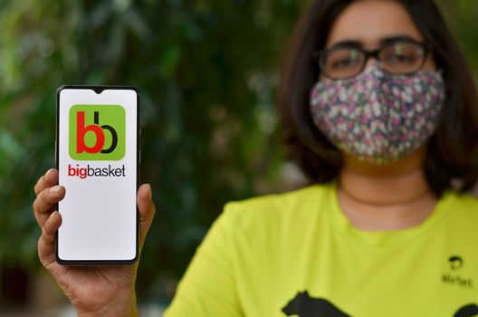 New Delhi, India, 2020. Girl wearing mask show Big Basket app on mobile under home quarantine during Corona virus (Covid-19) disease pandemic. Only delivery of essentials are allowed during lockdown