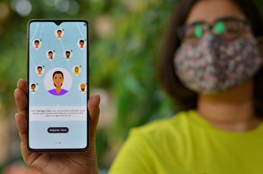 Delhi, India, 2020. Girl wearing mask showing Aarogya Setu app on mobile phone under home quarantine. It is launched by Indian govt for prevention & testing of Corona Virus (Covid-19) disease pandemic
