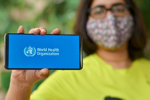 Geneva, Switzerland,2020. Girl wearing mask showing on her mobile phone screen, World health organization (WHO) landing page & emblem.Specialized agency of United Nations responsible for public health