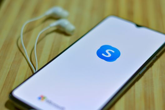 London, UK, 2020. Flat lay of Skype app icon on the smartphone screen on display and a pair of earphones on a wooden board background. SKype is a popular video call app