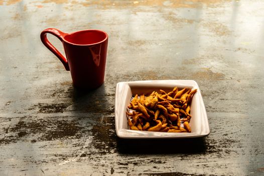 Close up Red Color Coffee mug and white plate full of Namkeen Bhujia snack cookies on old rustic floor. Food and drink background. Morning sunlight coming from window.